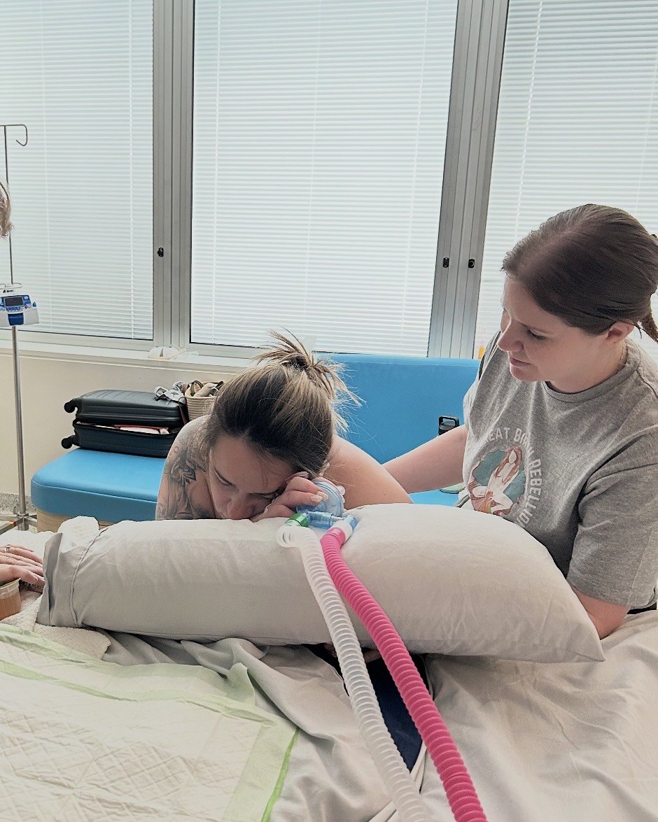 A labouring woman leaning forward over a pillow while her support person sits beside her